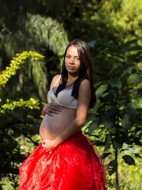 Gestante posa de bustiê de perolas e grande saia vermelha, no parque, com vegetação verde ao fundo'