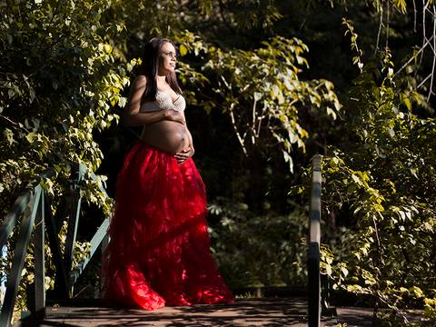 Gestante posa de bustiê de perolas e grande saia vermelha, posa na ponte estilo japonês, com vegetação verde ao fundo. Acaricia a barriga e olha para horizonte'