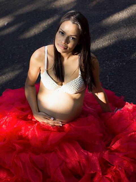 Gestante posa de bustiê de perolas e grande saia vermelha, no parque, acariciando a barriga e olhando para câmera'