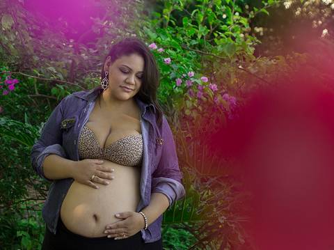 Gestante posa no parque, em meio a vegetação, com flores rosas emoldurando a imagem'
