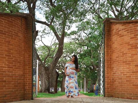 Gestante posa de vestido estampado, destacando a barriga, em frente a grande portão do parque'