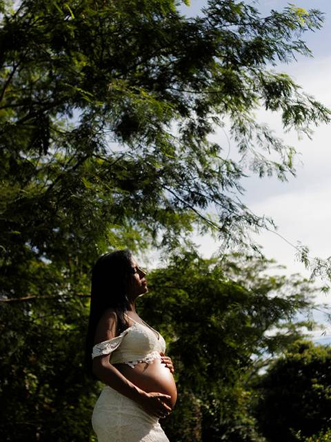 Gestante posa de roupa branca, destacando a barriga, em frente a grande e alta vegetação. o sol emoldura seu rosto e barriga'