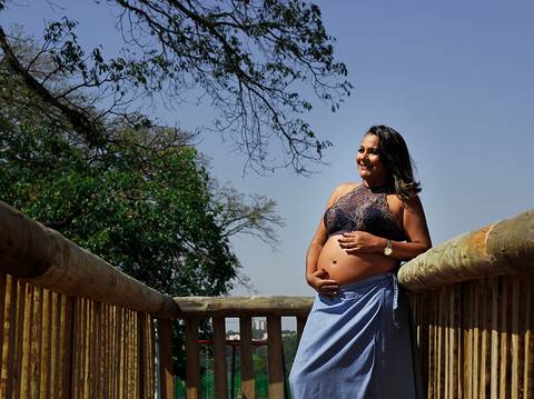 Gestante posa na casinha de madeira, sob o sol, olhando para o horizonte, e acariciando a barriga'