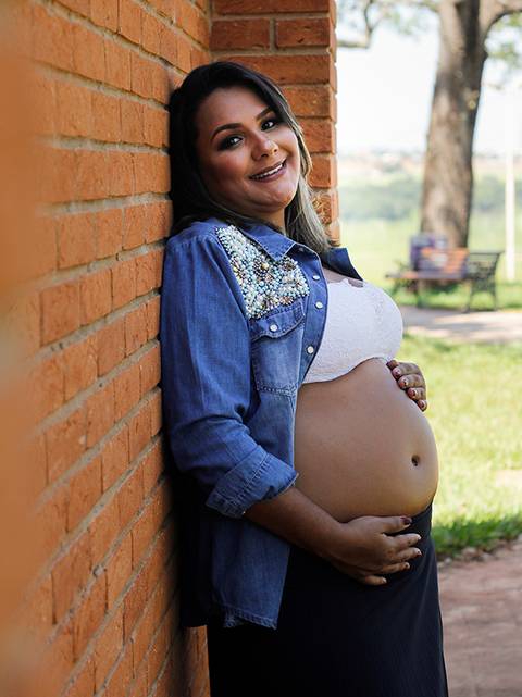 Gestante posa de lado na parede de tijolos. De camisa jeans, olha para câmera e acaricia a barria'