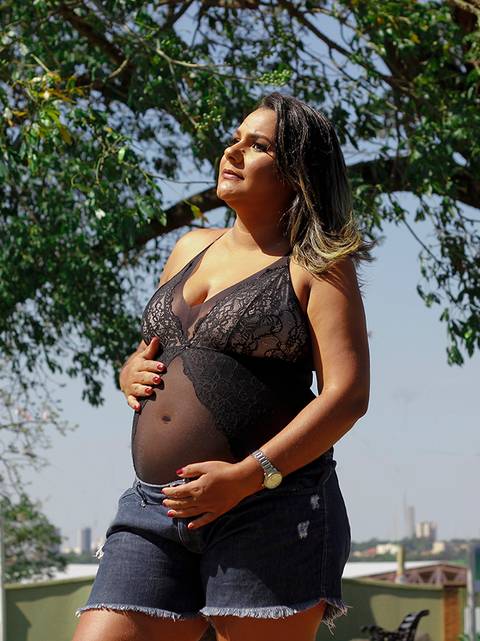 Gestante posa no parque, sob o sol, olhando para o horizonte, e acariciando a barriga'