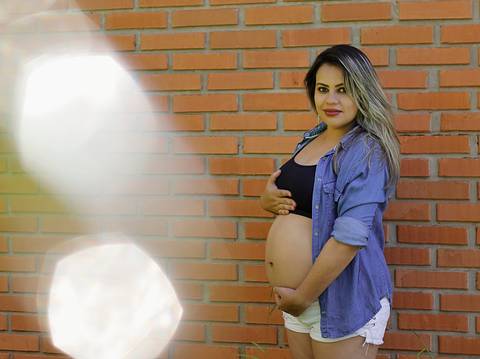 Gestante posa na parede de tijolos, de top e camisa aberta. Olha para câmera e destaca a barriga. Reflexo faz detalhe na foto.'