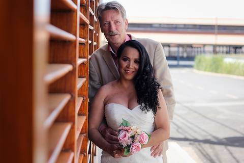 Noivo abraça noiva, a ambos posam para câmera, em um parede de madeira'