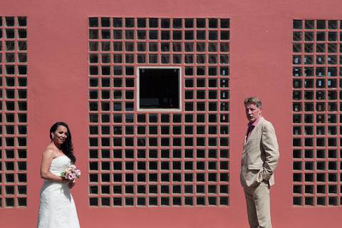 Noivos, em frente a parede vermelha com ladrilhos de vidro, de frente um para o outro, porem olhando para câmera'