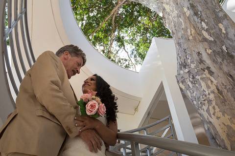 Noivos posam na escada abraçados se olhando. Foto de baixo para cima com copa de uma grande árvore ao fundo'