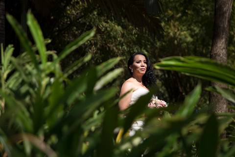 Noiva posa em meio a vegetação, olhando ao longe. Plantas verdes em desfoque no primeiro plano emolduram a noiva'