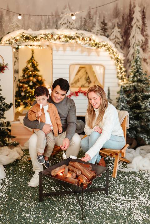 família branca Pai, Mãe e Filho de aproximadamente 3 anos de idade, sentados em cadeiras de acampamento segurando palitos de marshmallow em frente a uma fogueira realista. Trailer ao fundo com elementos natalinos e arvores de natal.'