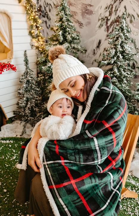 mãe e filha com touca de neve branca com pompom abraçadas em uma cadeira de acampamento com coberto verde xadrez em volta. ao fundo, cenário Natalino com uma parte de uma roda aparecendo.'