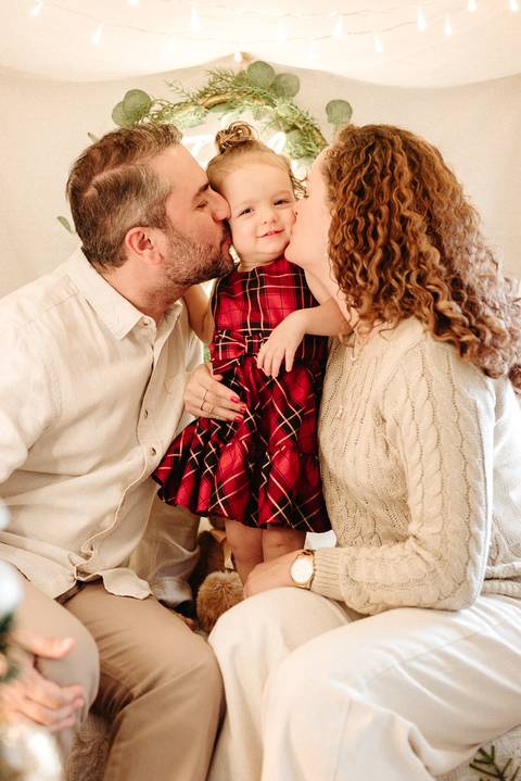pai e mãe dando um beijo um de cada lado em sua filha que veste um vestido xadrez vermelho e tem 1 ano de idade'