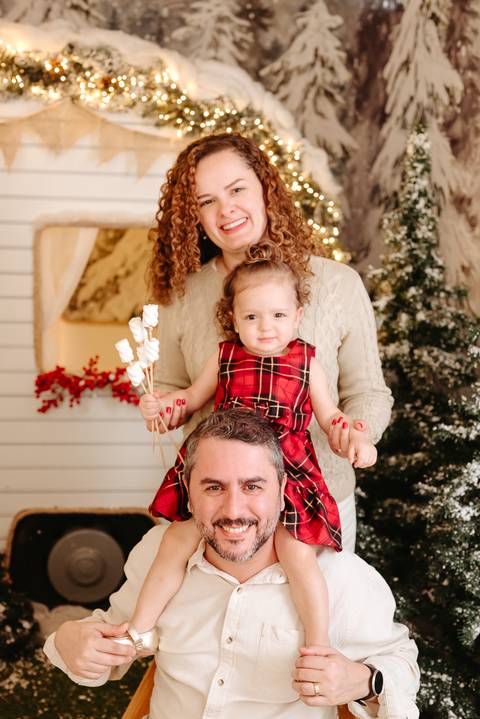 mãe de pé, filha que veste vestido xadrez vermelho sentada no ombro do pai que esta sentado em uma cadeira'