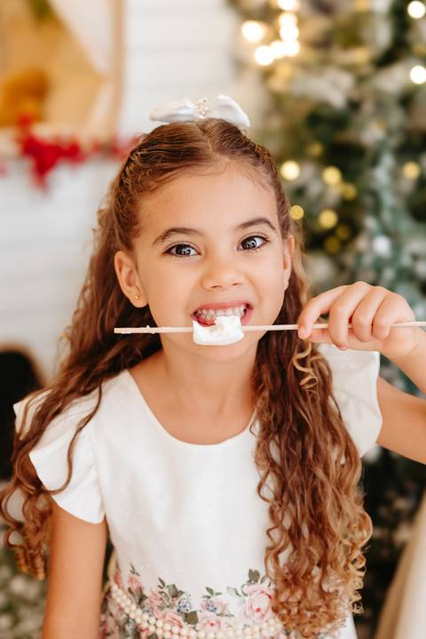 menina branca com cabelos cor Loiro escuro segurando um palito de espeto com marshmallow. ela esta mordendo o doce e olhando diretamente para a câmera. ao fundo, uma parte do trailer com desfoque de luzes de natal. cenário natalino.'