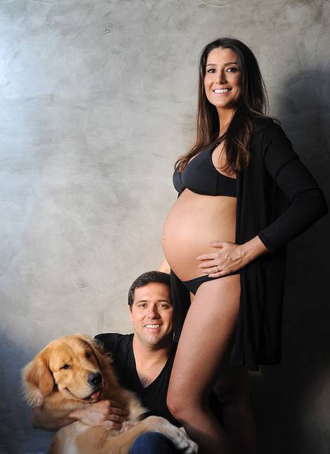 Casal posando com seu pet, homem sentado abraçado em seu pet Golden retriever, mulher grávida com uma das mãos na cintura, posicionada de lado, ambos sorrindo.'