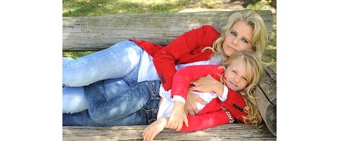Mãe e Filha deitadas em um banco de madeira rústico em um parque, ambas usando calça jeans e casaco vermelho, loiras com olhos claros.'