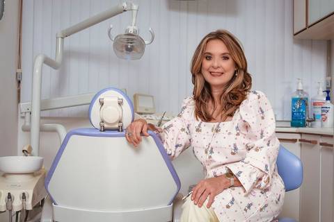 ensaio de marca pessoal da dentista em seu consultório, ela está sentada e apoia uma das mãos na cadeira de atendimento. Ela sorri levemente para a camera transmitindo autoridade.'