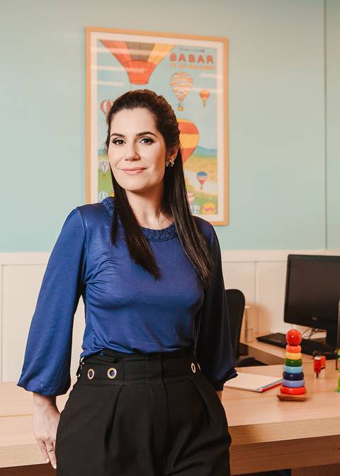 ensaio de marca pessoal em consultório médico em Niterói e Rio de Janeiro de médica pediatra ela olha para a camera em pé em frente a sua mesa de trabalho. Ela está com um leve sorriso transmitindo autoridade.'