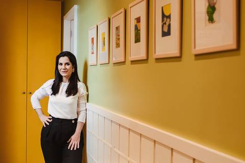  fotografia de retrato para médicos Niterói e rio de janeiro no consultório da pediatra que veste blusa off white e calça social preta. Ela está encostada em uma parede verde e uma linha quadros na parede guiam o olhar até ela.'