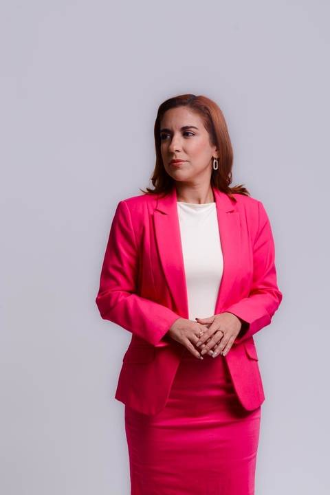 retrato profissional em estúdio de mulher empreendedora do ramos de maquiagem e beleza. Ela veste blaser rosa, saia rosa e blusa branca. Ela está olhando para o lado direito com leve sorriso, mãos sobrepostas demonstrando autoridade.'