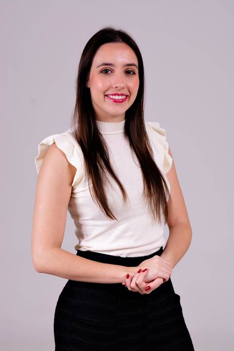 Retrato corporativo feminino de médica sorrindo em estúdio, com blusa off-white de babados e calça preta, postura confiante e profissional.'