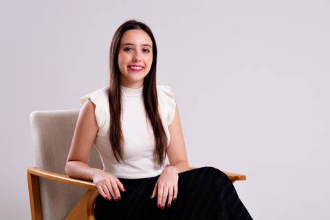 Ensaio corporativo feminino de empreededora sentada em uma cadeira de design moderno em um estúdio. A mulher está sorrindo, vestindo blusa off-white e calça preta, transmitindo confiança e acessibilidade. A camera está mais distante para ver toda a cena'