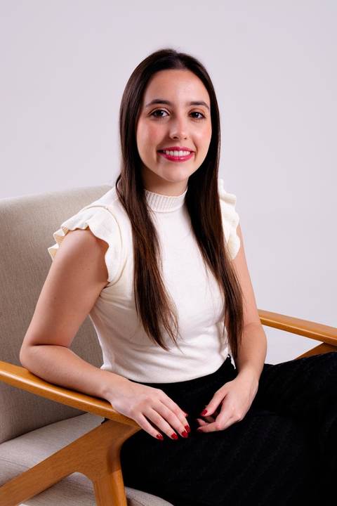 Ensaio corporativo feminino de médica sentada em uma cadeira de design moderno em um estúdio. A mulher está sorrindo, vestindo blusa off-white e calça preta, transmitindo confiança e acessibilidade.'