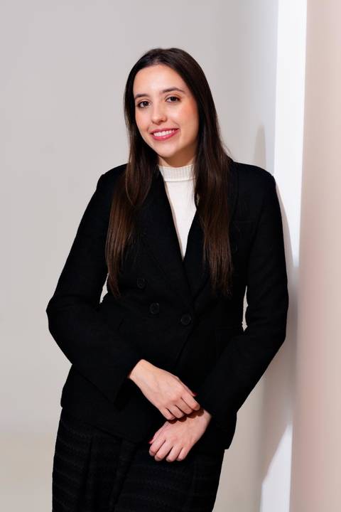 Retrato corporativo elegante de executiva em blazer preto, sorrindo, encostada em uma parede clara. A pose é moderna e acessível, ideal para consultoria.'