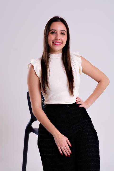 Retrato corporativo de empreendedora/consultora com pose confiante e mão na cintura, sorrindo e apoiada em uma cadeira preta. A foto em estúdio, com blusa clara e calça escura, transmite acessibilidade e determinação.'