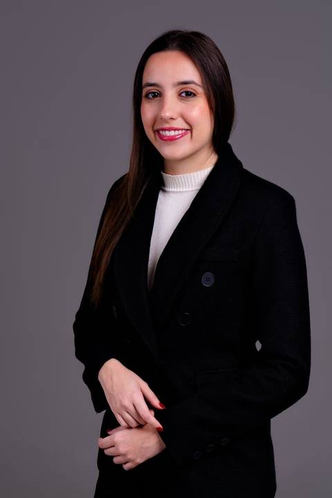 Foto profissional de meio corpo de mulher de negócios/executiva em blazer preto, com as mãos unidas. O fundo escuro e a pose reforçam a seriedade e o profissionalismo.'