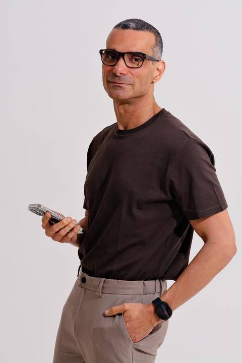 fotografia de marca pessoal de médico em estúdio fotográfico com luz suave. O médico está de pé olhando para a camera e segurando seu celular. Ele veste camisa básica marrom e calça sarja cinza.'