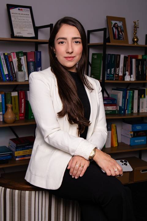 advogada em seu escritório sentada em uma cadeira e ao fundo estantes com livros jurídicos'