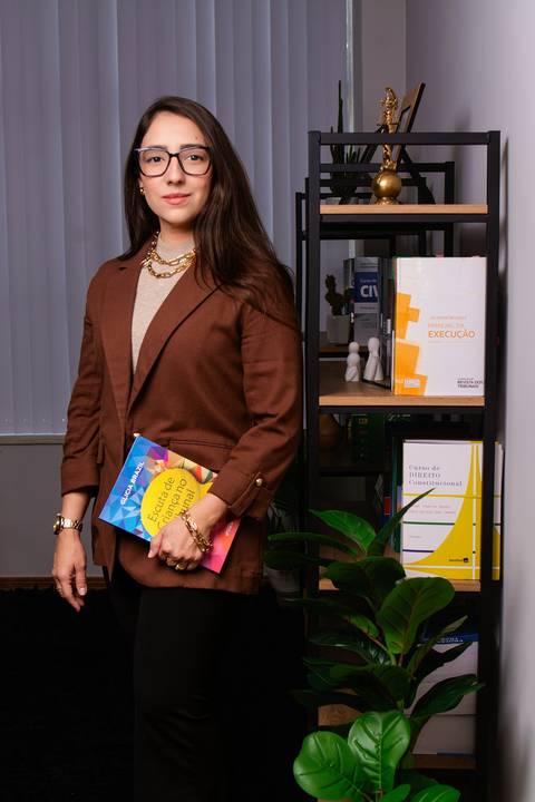 advogada ao lado de uma estante com livros em seu escritório segurando um livro ela olha diretamente para a camera'