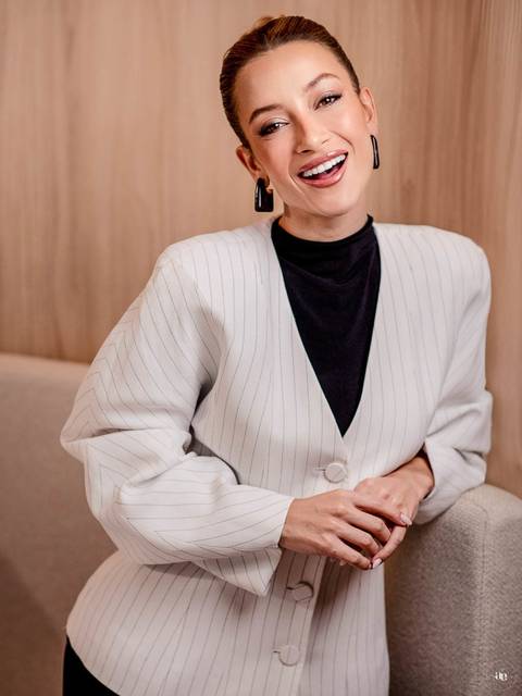 Retrato corporativo de Katiane Soares em blazer branco com risca de giz, sentada em ambiente de tons neutros, com postura elegante e linguagem corporal que transmite segurança profissional.'