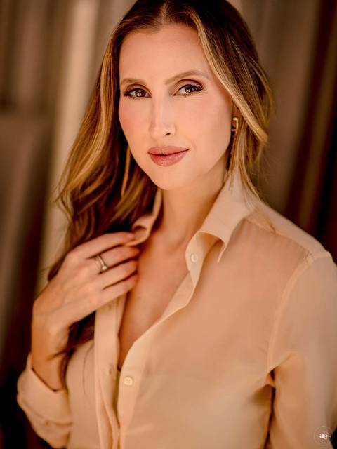 Fotografia profissional da estilista Paloma com camisa nude, expressão serena e mão próxima ao colo, destacando a naturalidade e a elegância do retrato.'
