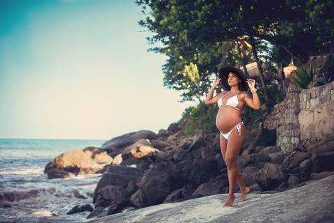 Book de gestante externo usando biquíni branco e chapéu preto e fazendo pose nas pontas dos pés-Prainha/RJ'