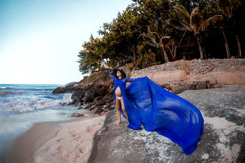 Gestante com vestido azul esvoaçante em cima de pedras em seu ensaio gestante-Prainha/RJ'