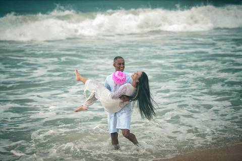 Ensaio casal noivo segurando noiva na colo enquanto caminham na praia'