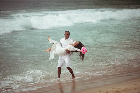 Casal caminhando na areia em praia, noivo segurando ela no colo'