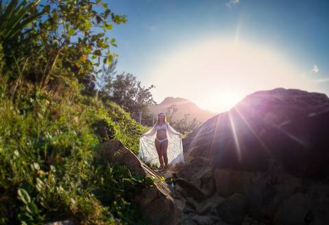 Gestante de braços abertos em cima da pedra ao fundo raios de sol  em seu ensaio fotográfico na prainha R-J'