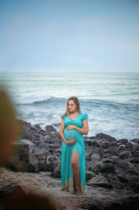 gestante segurando barriga em seu book fotográfico externo ao fundo pedras e mar na prainha/RJ'