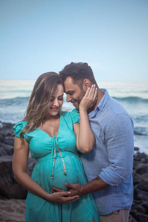 carinho entre papai e gestante em ensaio externo na prainha ao fundo mar e pedras'