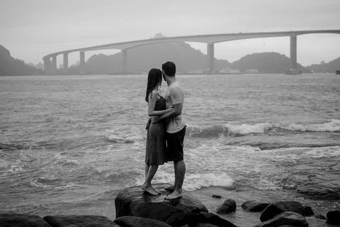 Retrato preto e branco do casal olhando para terceira ponte de vitória, ES. Casal em pé na pedra, próximo ao mar.'