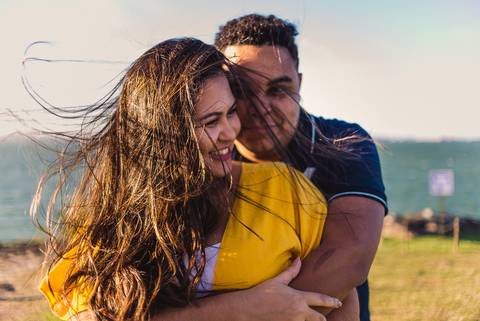 Fotografia externa de ensaio pré casamento. Casal abraçado. Cabelo da noiva embaraçado e voando devido a grande ventania no local.'