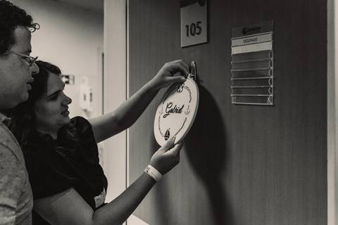 Casal colocando plaquinha na porta do quarto para receber o bebê após o parto. Plaquinha redonda pregada na porta'