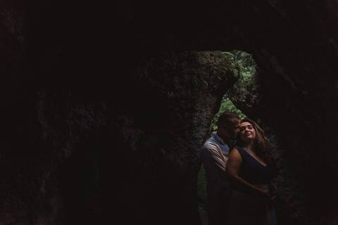 Fotografia do casal dentro de um rochedo e com uma luz entrando por uma fresta. Casal com olhos fechados.'