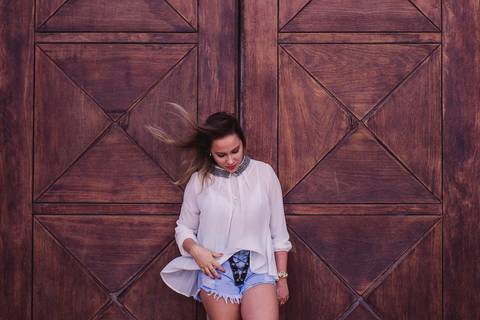 Fotografia do ensaio fotográfico pessoal, realizado na Ilha do Frade em Vitória - ES. Modelo em pé, olhando para baixo, com os cabelos ao vento em frente uma porta de madeira.'