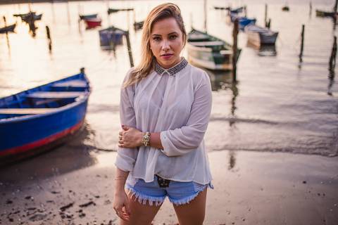 Retrato, modelo encarando no olhar. Em pé na praia com embarcações ao fundo da composição da foto.'