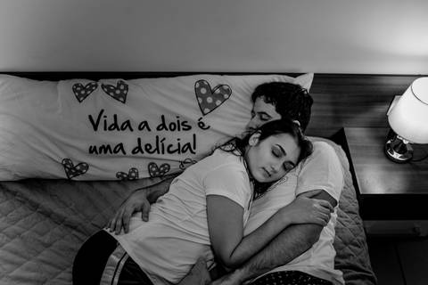 Fotografia - Ensaio de casal. Esposa deitada no peito do esposo. Este que está deitado com a cabeça sobre o travesseio que leva a frase: